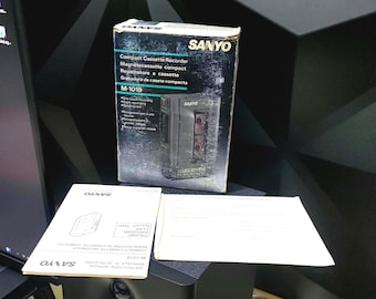 May include: A vintage Sanyo M-1019 compact cassette recorder in its original box. The box is black and blue with the Sanyo logo and product information. The image also shows the instruction manual and other paperwork.