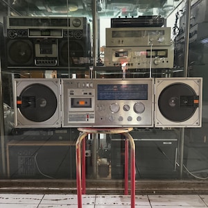 May include: A silver Sony portable stereo system featuring a central cassette deck, a radio tuner with a blue display, and two detachable side speakers. The unit has multiple circular control knobs, rectangular buttons, and a top-mounted handle.