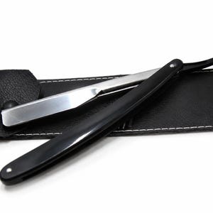 May include: A straight razor with a metallic silver blade and a smooth black handle, positioned diagonally across a black faux leather protective sleeve featuring white stitching along the edges and a single snap button closure.