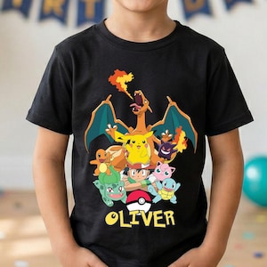 May include: Black t-shirt featuring a colorful graphic of various cartoon characters, including a dragon, Pikachu, and others, with the name "OLIVER" printed below. The shirt is worn by a smiling child.