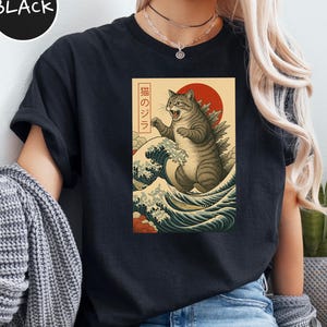 May include: Black t-shirt featuring a graphic of a cat-like monster in a Japanese art style, with a red sun and ocean waves. The text "猫のジラ" is in a rectangular frame. The word "BLACK" is in a black circle in the upper left.