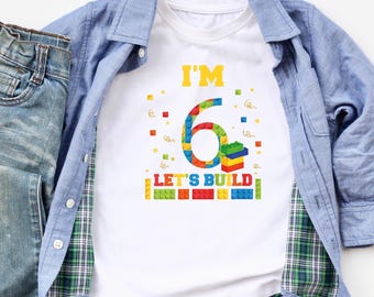 May include: White t-shirt with a colorful number six made of toy building blocks. The text "I'm 6 Let's Build" is printed on the shirt.