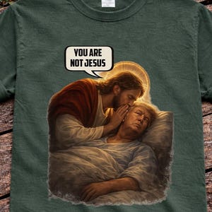 May include: Dark green t-shirt featuring a humorous graphic. The graphic depicts a religious scene with the text "YOU ARE NOT JESUS" in a speech bubble. The image shows a man whispering to another man.