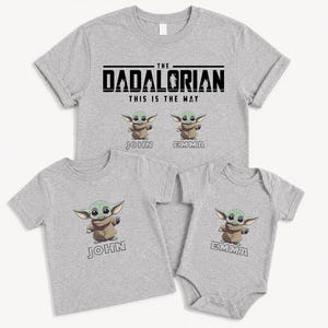 K&ouml;nnte beinhalten: Vier hellgraue T-Shirts und ein Body. Das gr&ouml;&szlig;te Shirt tr&auml;gt die Aufschrift "THE DADALORIAN THIS IS THE WAY" mit Cartoon-Bildern eines gr&uuml;nen Aliens. Die anderen Shirts und der Body zeigen den Alien und Namen.