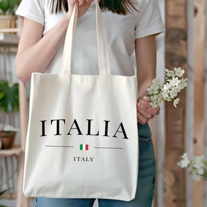 Puede incluir: Bolso tote de color crema con la palabra "ITALIA" en negro, encima de la palabra "ITALY" y la bandera italiana. El bolso lo sostiene una persona que lleva una camiseta blanca y vaqueros azules. Tambi&eacute;n se sostiene un peque&ntilde;o ramo de flores blancas.