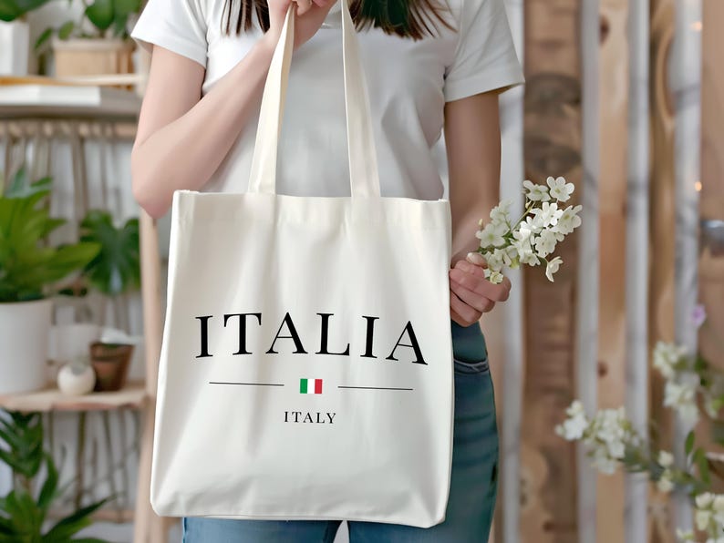 Puede incluir: Bolso tote de color crema con la palabra "ITALIA" en negro, encima de la palabra "ITALY" y la bandera italiana. El bolso lo sostiene una persona que lleva una camiseta blanca y vaqueros azules. Tambi&eacute;n se sostiene un peque&ntilde;o ramo de flores blancas.