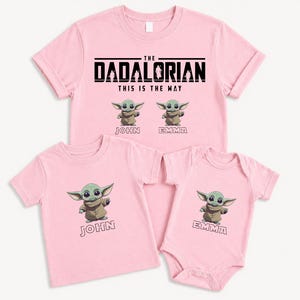K&ouml;nnte beinhalten: Rosafarbene T-Shirts und ein Body mit einer Zeichentrickfigur. Die Shirts tragen den Text "THE DADALORIAN THIS IS THE WAY" und das Bild der Figur. Die Shirts sind mit "JOHN" und "EMMA" beschriftet.