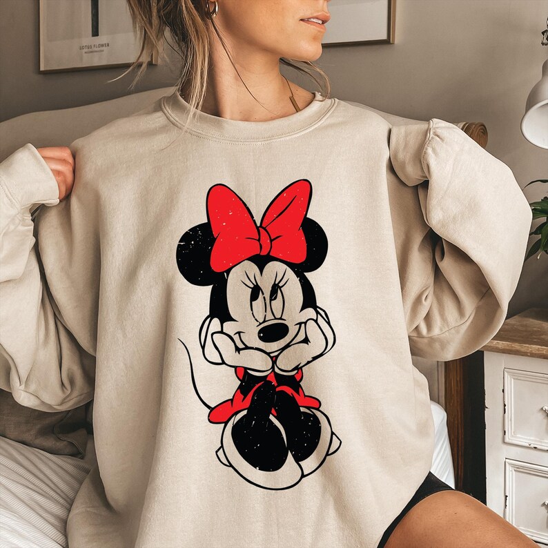 May include: Beige sweatshirt featuring a graphic of Minnie Mouse with a red bow and dress. The black and white illustration shows Minnie Mouse with her hands under her chin. The sweatshirt has long sleeves and a crew neck.