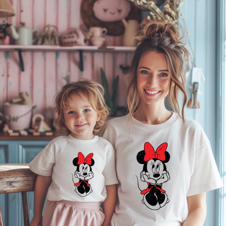 May include: Two people wearing matching off-white t-shirts featuring a Minnie Mouse graphic. The graphic shows Minnie Mouse with a red bow and dress. The shirts are worn by a woman and a young child, both smiling at the camera.