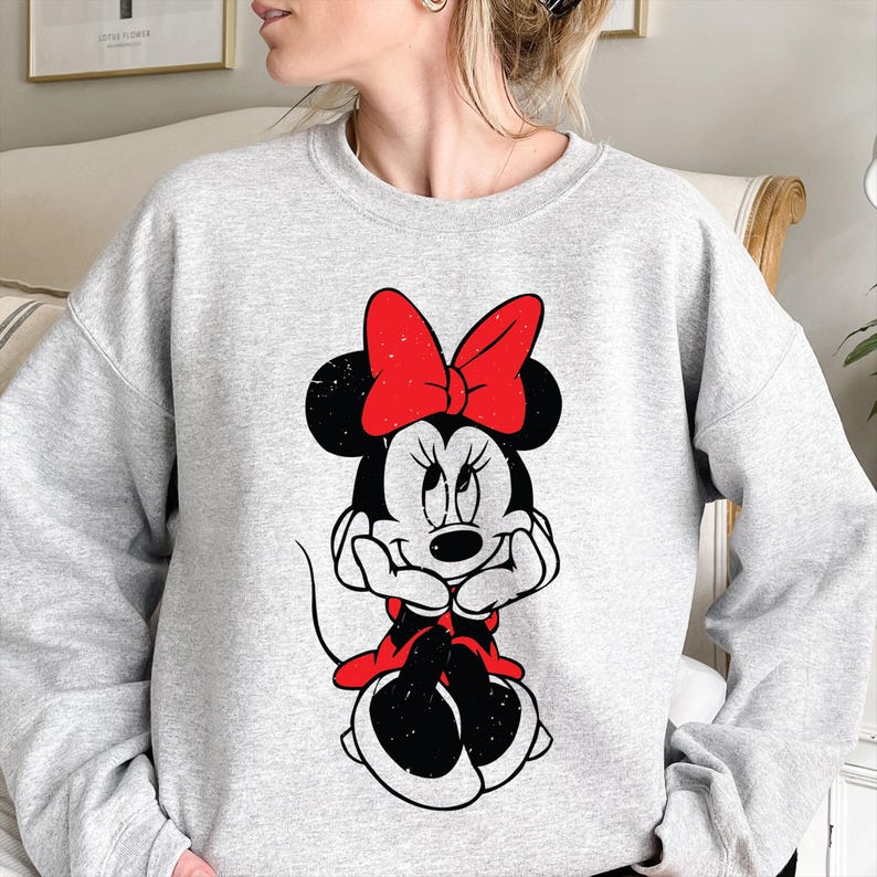 May include: A light gray sweatshirt featuring a black and white illustration of Minnie Mouse. Minnie Mouse is wearing a red bow and a red dress. The graphic is centered on the front of the sweatshirt.