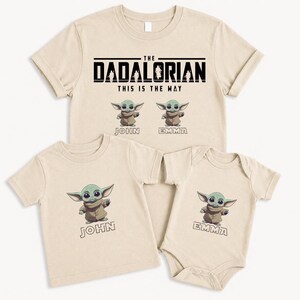 K&ouml;nnte beinhalten: Beige T-Shirts und ein Body mit einer Cartoonfigur. Das Erwachsenen-Shirt tr&auml;gt die Aufschrift "The Dadalorian" mit "This is the Way" darunter. Die Kinderhemden haben das Bild der Figur und die Namen "John" und "Emma".