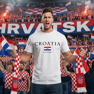 May include: A white t-shirt with "CROATIA" printed in black, a Croatian flag graphic, and the word "CUSTOM" in black. The image shows a person holding a Croatian flag and scarf in a stadium filled with fans.