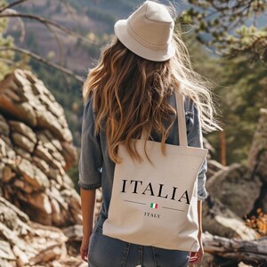 Puede incluir: Un bolso tote beige con la palabra "ITALIA" impresa en negro, junto con la bandera italiana y la palabra "ITALY" debajo. El bolso lo lleva una persona con sombrero y chaqueta vaquera, con un fondo monta&ntilde;oso.
