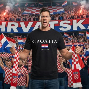 May include: A black t-shirt with "CROATIA" printed in white, featuring the Croatian flag. The image shows a person holding a scarf and flags with the Croatian coat of arms, in a stadium filled with fans.