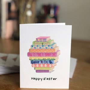 May include: A white card with a stack of colorful books with titles related to Easter. The books are stacked on top of each other and the top book has the title "The Egg Hunt". The card has the text "Happy Easter" at the bottom.