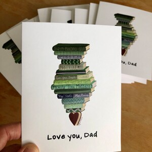 May include: A white card with a stack of green and blue books with titles like "Building Stuff", "Awesome", "Barbeque", "Save Your People", "Family Time Matters", "My Dad", "My Prince", and "Home-Fishing". A brown heart is at the bottom of the stack. The card says "Love you, Dad" in black handwriting.