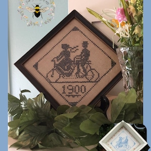 May include: A framed cross-stitch design of a couple riding a bicycle together, with the year 1900 stitched below. The frame is dark brown wood and the design is on a tan fabric. The image is titled "Bicycle Built for Two Silhouette".