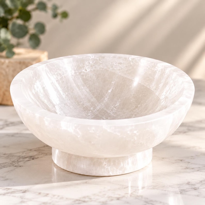 Puede incluir: Un cuenco de piedra pulida de color blanco transl&uacute;cido con un interior liso y redondeado y una base circular. El material presenta texturas cristalinas naturales y vetas sutiles en toda la superficie, lo que crea un aspecto brillante y reflectante.