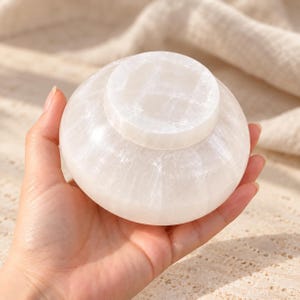 Puede incluir: Un cuenco peque&ntilde;o y redondo de selenita blanca con una superficie lisa y pulida y una textura cristalina transl&uacute;cida. El cuenco presenta un cuerpo ancho y curvado con una base circular elevada que muestra el aspecto fibroso.