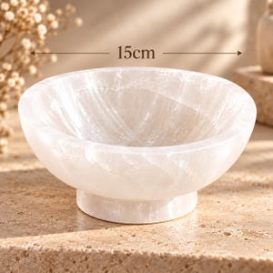 Puede incluir: Un cuenco de piedra pulida, blanco y transl&uacute;cido, con un borde redondeado y liso y una base de pedestal resistente. El cuenco presenta una textura cristalina fibrosa natural y mide 15 cm de di&aacute;metro, seg&uacute;n indica el texto de la imagen.