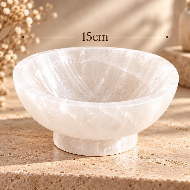 Puede incluir: Un cuenco de piedra pulida, blanco y transl&uacute;cido, con un borde redondeado y liso y una base de pedestal resistente. El cuenco presenta una textura cristalina fibrosa natural y mide 15 cm de di&aacute;metro, seg&uacute;n indica el texto de la imagen.