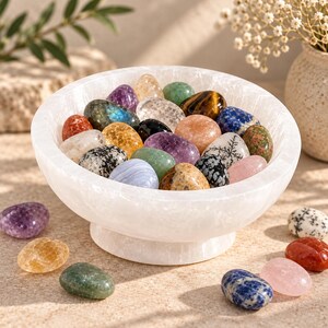 Puede incluir: Un cuenco de pedestal de selenita blanca lleno de una variedad de piedras preciosas pulidas y multicolores, incluyendo amatista, ojo de tigre, sodalita, cuarzo rosa y labradorita, con varias piedras sueltas esparcidas alrededor de la base.