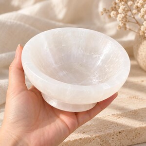 Puede incluir: Un cuenco peque&ntilde;o y redondo de selenita blanca con un acabado liso y pulido y una textura cristalina transl&uacute;cida. El cuenco presenta una base ancha y abierta y una base circular resistente, mostrando vetas minerales naturales.