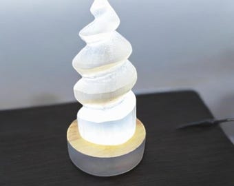 May include: A white crystal sculpture in the shape of a spiral, illuminated from within, sits on a wooden base. The sculpture is on a dark surface.