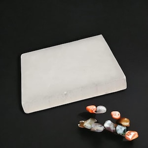 May include: A white rectangular slab of selenite crystal, a type of gypsum, on a black background. Several small, colourful stones are scattered around the slab.