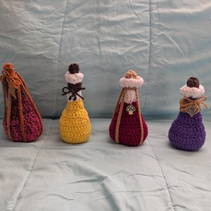 Op de afbeelding: Vier handgemaakte gehaakte toverdrankflessen in rood, geel en paars. Elke fles heeft een uniek ontwerp, inclusief decoratieve doppen en versieringen. De flessen zijn gerangschikt op een lichtblauwe ondergrond.