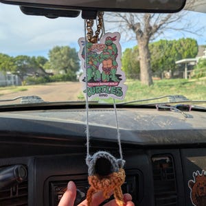 Puede incluir: Un ambientador de Teenage Mutant Ninja Turtles y un pequeño objeto de ganchillo colgando del espejo retrovisor de un coche. El ambientador muestra a las Tortugas con pizza. El objeto de ganchillo es gris y marrón.