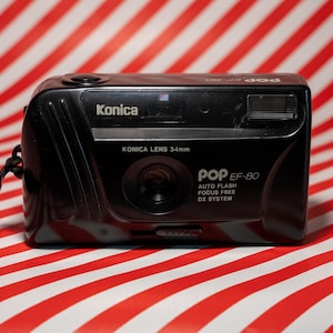 Peut inclure: Un appareil photo argentique noir Konica Pop EF-80 35 mm avec flash intégré. L'appareil photo est muni d'une sangle noire et repose sur une surface à rayures rouges et blanches.