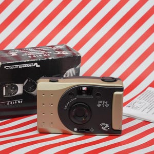 Peut inclure: Un appareil photo compact de couleur dorée avec un objectif noir et le texte "PN 919" sur le devant. L'appareil photo est posé sur une surface à rayures rouges et blanches.