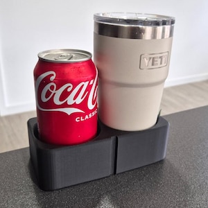 Puede incluir: Una lata roja de Coca-Cola y un vaso Yeti beige se encuentran en un portavasos doble negro. La lata de Coca-Cola tiene letras blancas y una tapa plateada. El vaso Yeti tiene un borde plateado y el logotipo de Yeti.