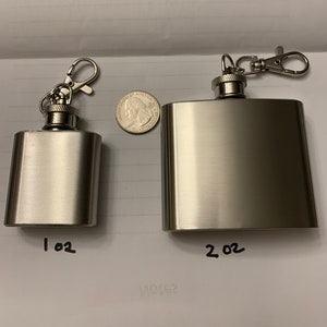Op de afbeelding: Twee roestvrijstalen hipflasks met bevestigde sleutelhangers. De kleinere fles is gemarkeerd met "1 oz" en de grotere fles is gemarkeerd met "2 oz".