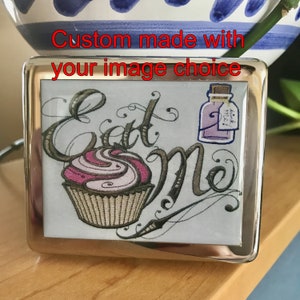 Può includere: Un compatto in metallo argentato con un'immagine colorata di un cupcake e una bottiglia con il testo "Eat Me" e "Drink Me!" all'interno.