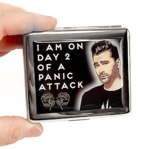 May include: A silver cigarette case with a black and white image of a man with the text "I am on day 2 of a panic attack" and two roses.
