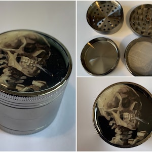 May include: A silver metal grinder with a skull and cigarette design on the top. The grinder has four parts: a top lid, a grinding chamber, a screen, and a bottom collection tray.