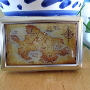 May include: A silver metal box with a hinged lid. The lid features a colourful map with the words "Skull Rock", "Mermaid Lagoon", "Cannibal Cove", and "Hangman's Tree".
