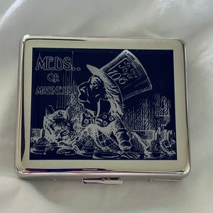 May include: A silver cigarette case with a black and white image of the Mad Hatter from Alice in Wonderland. The image has the text "MEDS... OR MADNESS!" and "In this style 10/6".