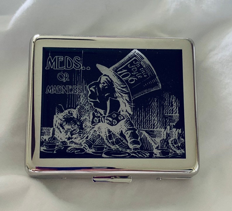 May include: A silver cigarette case with a black and white image of the Mad Hatter from Alice in Wonderland. The image has the text "MEDS... OR MADNESS!" and "In this style 10/6".