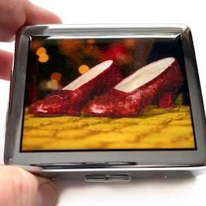 Op de afbeelding: Een zilveren compact met een afbeelding van een rode robijn slipper. De afbeelding toont de slippers op een gele bakstenen weg.