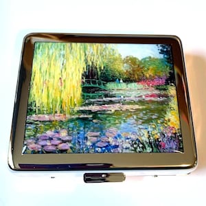May include: A silver metal cigarette case featuring a colorful impressionist painting of a pond with water lilies, a small bridge, and weeping willow trees. The artwork is framed by a shiny silver border.