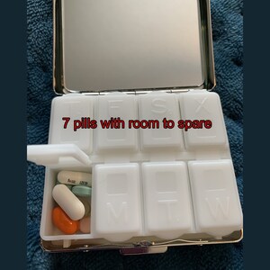 May include: A white pill organizer with a mirror lid. The organizer has seven compartments labeled with the days of the week. The compartment for Monday is open and contains seven pills. The text "7 pills with room to spare" is written on the organizer.