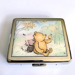 Pode incluir: Um estojo de cigarros de metal prateado com uma ilustração do Winnie the Pooh e do Porquinho. A ilustração mostra o Winnie the Pooh e o Porquinho sentados em uma clareira na floresta com árvores e flores.