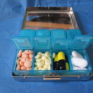 Puede incluir: Un organizador de pastillas de pl&aacute;stico azul con siete compartimentos. Los compartimentos contienen varias pastillas, incluyendo pastillas amarillas, naranjas y blancas, as&iacute; como dos c&aacute;psulas negras y amarillas.
