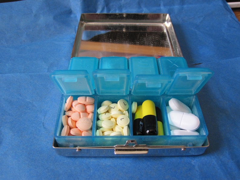 Puede incluir: Un organizador de pastillas de pl&aacute;stico azul con siete compartimentos. Los compartimentos contienen varias pastillas, incluyendo pastillas amarillas, naranjas y blancas, as&iacute; como dos c&aacute;psulas negras y amarillas.