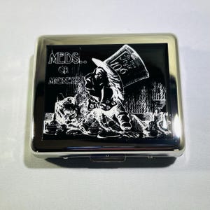 May include: A silver cigarette case with a black background and a white image of the Mad Hatter from Alice in Wonderland. The text "MEDS... OR MADNESS" is printed on the case.