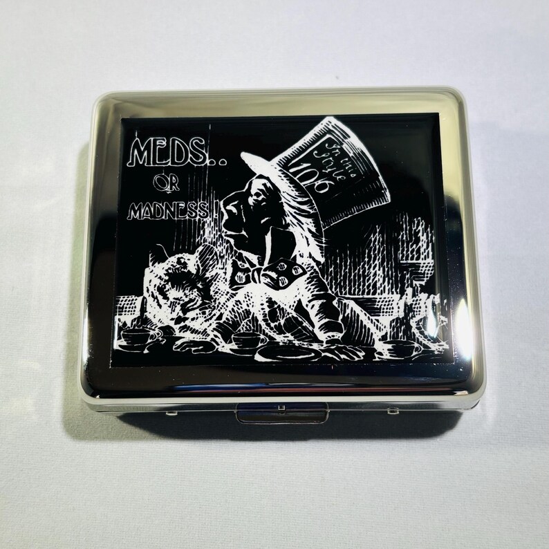 May include: A silver cigarette case with a black background and a white image of the Mad Hatter from Alice in Wonderland. The text "MEDS... OR MADNESS" is printed on the case.