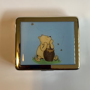 May include: A silver metal Winnie the Pooh trinket box with a blue background and a picture of Winnie the Pooh sitting with a pot of honey.  The text "HUNNY" is on the pot of honey.  There are bees flying around Winnie the Pooh.